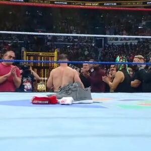 WWE superstar john cena kneels in the center of the ring with his shirt and armband placed on the mat, as fellow wrestlers and a packed arena stand and applaud in an emotional tribute moment.