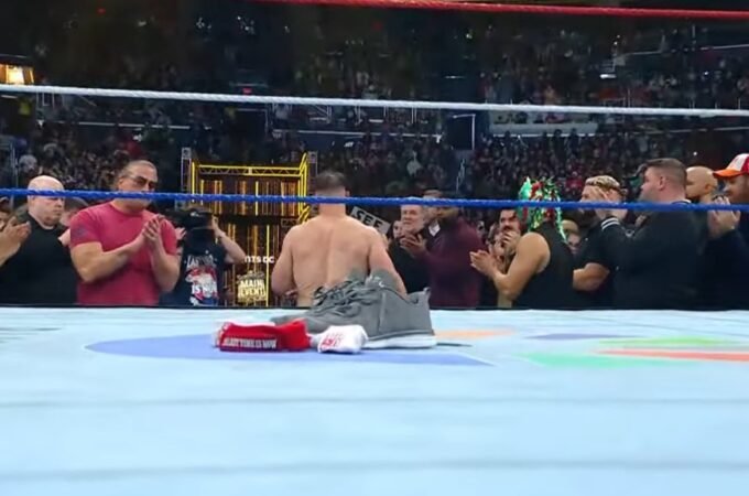 WWE superstar john cena kneels in the center of the ring with his shirt and armband placed on the mat, as fellow wrestlers and a packed arena stand and applaud in an emotional tribute moment.