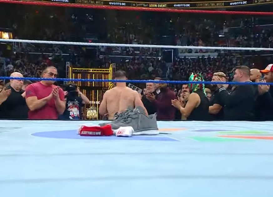 WWE superstar john cena kneels in the center of the ring with his shirt and armband placed on the mat, as fellow wrestlers and a packed arena stand and applaud in an emotional tribute moment.